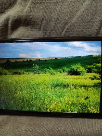 The image displays a screen showing a rolling green landscape with a bright blue sky dotted with white clouds. In the foreground, tall green grass is dominant, with some smaller plants and flowers visible. Further back, there are undulating green hills interspersed with trees and shrubs. The horizon is marked by fields of a lighter brown hue, suggesting agricultural land. The scene appears serene and natural, with no people or animals present. The image is presented on a screen, as evidenced by the visible bezel and the texture of the surface beneath, which appears to be a coarse fabric. The lighting is bright, indicating daytime. There are some visual distortions on the screen, possibly due to damage or the angle of the photo. No specific landmarks or features are identifiable to pinpoint a location within Svoboda, Russia, but the landscape suggests a rural or semi-rural setting.