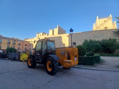A telehandler, identified by the "MAQUINZA" logo and model number "540-180", is parked on a paved surface in front of a stone wall. The telehandler is yellow and black. Behind the telehandler and wall, buildings with light-colored facades and windows are visible. The sky is clear and blue. The scene appears to be in an urban environment, possibly a public square or street in Ávila, Spain, given the architecture of the buildings and the style of the wall, which resembles the ancient city walls of Ávila. There are no people visible in the image. Several large trash bins are also present to the left of the telehandler.