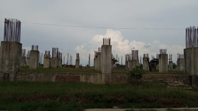 The image depicts an under-construction building site with numerous concrete pillars and exposed rebar, stretching across the frame. The scene is set outdoors under a cloudy sky, suggesting it might be late afternoon or early morning. The foreground is covered in lush green grass, with some areas showing overgrown vegetation. In the mid-ground, a low concrete wall runs horizontally, separating the grassy area from the elevated foundation of the building.
The main subjects are the architectural elements of the construction site. Many tall, cylindrical concrete pillars stand in rows, their tops adorned with bundles of vertical rebar, ready for the next stage of construction. Some of these pillars are connected by horizontal beams, also with exposed rebar. In the background, more pillars and construction scaffolding are visible, indicating a large-scale project. A few sparse trees and bushes are scattered throughout the site, adding a touch of nature to the otherwise industrial landscape. A single, small green leafy plant is visible to the right of the frame, contrasting with the concrete.
There are no people visible in the image. The overall mood is one of progress and development, but also a sense of unfinishedness. The cloudy sky and overcast lighting create a somewhat subdued atmosphere. There are no visible signs of text in the image. The location context provided is Keudeteupinpunti, Indonesia.