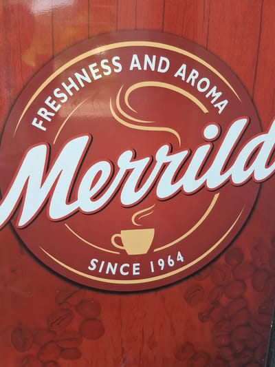 This is a close-up, eye-level photograph of a sign for "Merrild" coffee. The sign is predominantly red with a circular emblem in the center, which is also red and bordered by a thin yellow line. Within the emblem, white text reads "FRESHNESS AND AROMA" arched across the top. Below this, in large white script with a red outline, is the brand name "Merrild." Beneath "Merrild" is a stylized yellow image of a coffee cup with steam rising from it, and below that, white text reads "SINCE 1964." The background of the sign appears to be a wooden texture. The lower portion of the image shows a scattering of coffee beans, also in a reddish hue. There are no people or animals visible in the image. The location in Lēdmane, Latvia, is not discernible from the image itself.