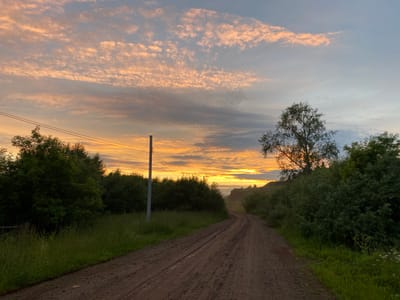The image shows a dirt road winding through a rural landscape at sunrise. The sky is filled with dramatic clouds painted in hues of orange, pink, and gray. On either side of the road are dense green trees and tall grass. A single utility pole with wires stands to the left of the road, and a large tree with silhouetted branches is visible on the right. The road appears to be slightly wet in places, with tire tracks visible. In the distance, a gentle hill rises, also covered in vegetation. The scene suggests a tranquil morning in a natural setting outside of the city of Baklushi, Russia.