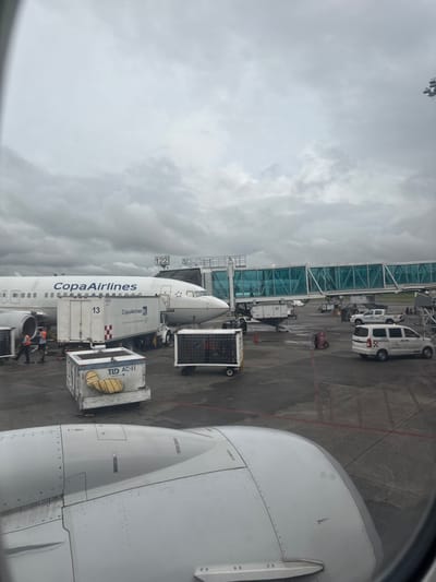 An airplane at an airport is shown from inside another plane. The view through the window includes the wing of the observer's aircraft. Adjacent is a Copa Airlines plane, docked at gate 122 with ground service equipment attached. Gray, overcast skies loom above the passenger boarding bridge. Multiple service vehicles are also visible on the tarmac.