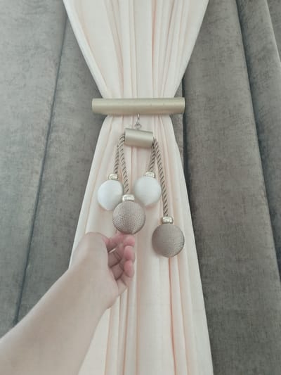 An arm is reaching out to touch the tassel of a curtain tie-back. The tie-back consists of a gold bar and a braided cord with three decorative balls: two white and one gold. The curtains are light pink and grey. The scene appears to be indoors, possibly a home, with no specific identifiable landmarks in Mady, Kyrgyzstan.
