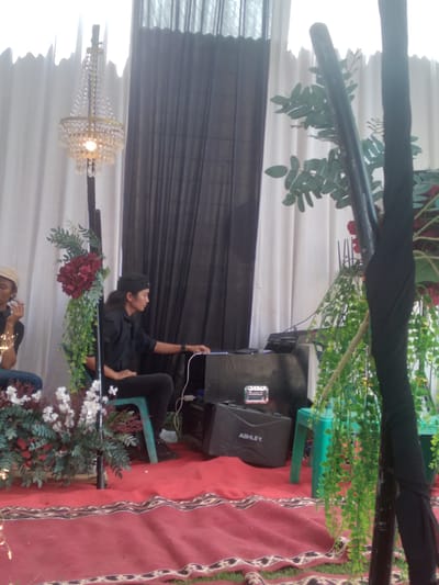 The image depicts an indoor or semi-outdoor event setting in Matangbaroh, Indonesia. The backdrop consists of floor-to-ceiling drapes: white pleated curtains on the left and black pleated curtains on the right. The floor is covered by a large red carpet with a patterned border in brown and white, laid over what appears to be grass.

Two men are present. On the far left, a man wearing a light-colored cap and dark shirt is partially visible, seated and holding a small object to his mouth. In the center-right, a man with long hair, wearing a black shirt, black pants, and a black head covering, is seated on a teal plastic stool. He is actively engaging with and operating electronic equipment, likely a sound mixer, placed on a dark surface in front of him. Cables are visible connecting to this equipment. Below the equipment, a black "ASHLEY" brand case with a silver handle rests on the carpet, alongside another dark case or speaker.

The scene is decorated with several items. A golden-toned chandelier with multiple tiers of crystal beads and a single glowing bulb is suspended from a dark pole on the left. This pole is also adorned with artificial floral arrangements, including dark red hydrangea-like flowers and green foliage. At the bottom left, additional white flowers and green leafy plants are arranged on the carpet. On the right, a thick, dark pole or structure, possibly wrapped in black fabric, is decorated with long strands of artificial green vines. A second teal plastic stool is partially visible on the far right.