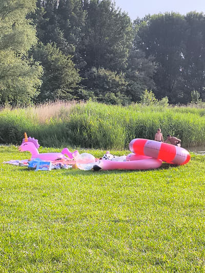 An outdoor scene on a sunny day in Broek op Langedijk, Netherlands, depicts a grassy area by a body of water, likely a canal or river, bordered by tall reeds and trees. In the foreground, an assortment of inflatable items is spread out on the grass, including a pink unicorn float, a circular pink float with a black and white cow print pattern, and a large red and white striped inflatable ring. A blue box is placed on a striped blanket. Further back, two shirtless individuals, appearing to be a man and a boy, are near the water's edge, with the man interacting with the boy or the water. The overall impression is a relaxed, recreational setting.