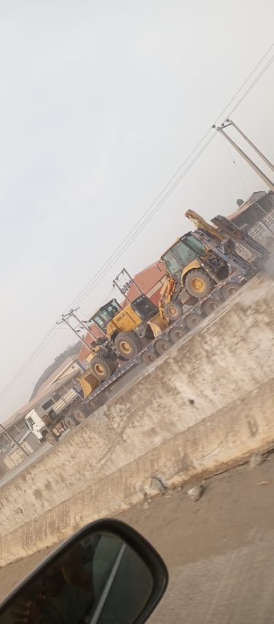 The image captures a roadside scene in Iddah, Nigeria, from the perspective of a moving vehicle, evidenced by a black side mirror visible in the lower left. Two heavy-duty trucks are transporting construction machinery on multi-axle flatbed trailers. The foremost truck is a white prime mover pulling a trailer laden with a yellow SBLG wheel loader, marked with "650087". Behind this, a second truck transports additional yellow construction equipment, including a machine with a visible boom and grapple attachment. Overhead utility poles with power lines are present. In the background, residential or commercial buildings with reddish-brown roofs are visible under a hazy, pale sky, suggesting dusty atmospheric conditions. A concrete barrier extends diagonally across the mid-ground, separating the viewer's immediate vicinity from the road where the trucks are traveling.