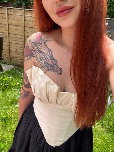 A medium shot captures a person with long, auburn hair and a dragon tattoo on their chest and arm, wearing a cream-colored corset top and a black skirt, standing outdoors in a grassy area with a wooden fence behind them. The person is not in Oldham, United Kingdom.