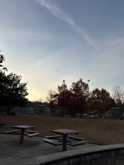 A park scene shows picnic tables on concrete near trees with changing leaves. In the background, cage-like structures stand amidst the trees, possibly baseball field netting. The sky transitions from a clear blue with a jet contrail to a lighter hue near the horizon, suggesting dawn or dusk. A concrete structure or ledge defines the edge of the park area.