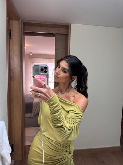 An adult female is depicted in a mirror selfie within an indoor environment. She is positioned centrally, facing slightly to her left, with her head turned to look at her reflection. She possesses dark hair, styled in a ponytail, and wears a light green, off-the-shoulder dress featuring ruched detailing on the long sleeves and a gathered, knotted element on the left side of the waist. She wears a thin, silver-toned necklace and two rings on her right hand: one with a prominent oval green stone on the ring finger and another with an abstract design on the middle finger. Her fingernails are painted a dark color.
The subject is holding a light-colored smartphone, likely an iPhone with three visible camera lenses, in her right hand. The phone is encased in a clear cover, to which a pink "pop-it" style fidget accessory is attached on the back. A white cable hangs from the phone, partially obscuring the dress.
The background, reflected in the mirror, reveals a portion of an interior room. A wooden door frame with a black hinge is visible on the left, suggesting an open doorway to an adjacent space. The reflected room contains a bed with light-colored bedding and a window with a light-toned blind or curtain. The walls are light-colored, appearing beige or off-white. Directly visible to the far left is a white towel hanging on a fixture. The visible flooring in the foreground and reflection appears to be wood. There are no discernible visual elements within the image to confirm the specific geographic location as San Luis Potosí City, Mexico.