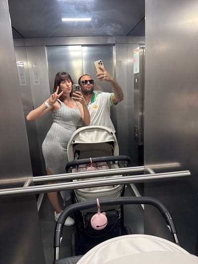 In an elevator interior, a man and a woman are visible in a mirror taking a selfie. The man, with a beard and wearing sunglasses, is holding a smartphone in his left hand. He is wearing a white t-shirt with green and yellow accents. The woman, wearing a grey dress, holds up two fingers in a peace sign. A stroller occupies the right side of the elevator. The elevator's metal interior is visible with control panels and signage on the walls. The location is in Pomezia, Italy, based on signage visible in the mirror.