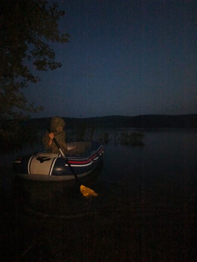 A person wearing a camouflage hooded jacket sits in an inflatable boat on a dark body of water. The boat has blue, white, and red stripes. The person is holding an oar, with the paddle visible. To the left of the boat, a tree branch with leaves hangs into the frame. In the background, a dark, undulating treeline is visible against a dark blue sky. The water appears still, with some indistinct vegetation or ripples in the middle ground. The scene is dimly lit, suggesting it is either dusk or dawn, or an overcast night.