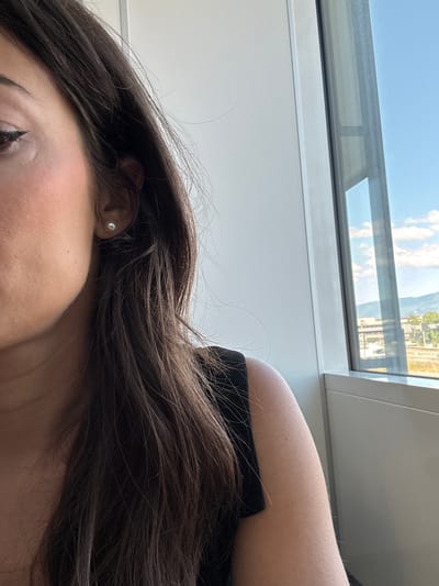 A medium shot shows the side profile of a woman with long, dark hair. She is wearing a black top and a small pearl earring. She is positioned in front of a large window that offers a view of a city skyline with buildings and mountains in the distance. The sky is blue with scattered clouds. The setting appears to be indoors, possibly an office or apartment, in Le Grand-Saconnex, Switzerland, given the window's view.