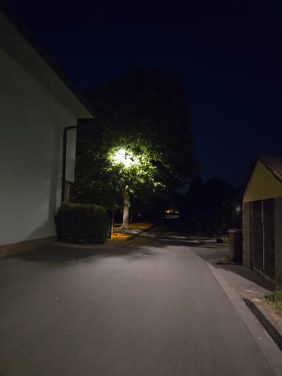 The image depicts a street scene at night in Kostelec na Hané, Czechia. The foreground features a paved road or pathway that curves to the right. To the left, a building with a light-colored facade and a dark gutter system is visible. Adjacent to the building, a dark green hedge or bush is present. A large tree with illuminated foliage, due to a nearby street lamp, is centrally located in the mid-ground. Beyond the tree, the scene transitions into darkness, with silhouetted trees and structures. On the right side of the image, a building with a yellowish facade and a dark roof is partially visible, alongside a dark-colored trash receptacle. The sky is a deep, dark blue, indicative of nighttime. There are no people or animals visible in the scene.
