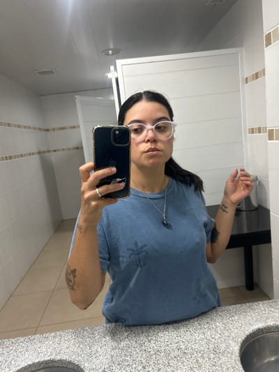 An adult individual is captured in a mirror selfie, standing upright and holding a smartphone to take the photo. The person has dark hair worn down, and wears light-framed glasses. Their expression is neutral.
They are wearing a blue, short-sleeved t-shirt with a subtle dark print on the front, which appears to be a botanical design, possibly a palm tree. A long silver chain necklace with a dark, possibly oval-shaped, pendant hangs around their neck. A silver ring is visible on their right ring finger. Several tattoos are visible on their arms; a small design on the right forearm and a larger, more intricate design on the upper left arm.
The smartphone used for the selfie is dark-colored, likely an iPhone, with a clear or light-colored protective case featuring gold or metallic edges. A white sticker or pop socket, depicting what looks like a cartoon character (possibly an animal with a bow), is affixed to the back of the phone case.
The setting appears to be a bathroom. A large mirror reflects the person and the surroundings. To the right of the person, a speckled grey and white granite-like countertop is visible, with the edge of a sink basin. The walls are white, featuring a horizontal strip of light brown and beige mosaic-like tiles near the top. The ceiling is white, and a light fixture or vent is visible in the reflection of the mirror. In the background reflection, there's also a white door or cabinet and another light fixture on the wall.