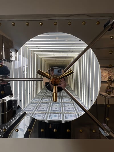 The image shows a close-up view of a large, circular vault door. The door is made of polished metal and features a prominent gold-colored handle. Inside the vault, which appears to be an infinity mirror installation, stacks of US dollar bills are arranged on shelves that recede into the distance. The walls of the vault are lined with vertical white lights, creating a tunnel effect. A reflection of a person holding a smartphone is visible in the vault door. In the background, outside the vault, a table with some decorative items is visible. The scene is lit by a combination of the internal vault lighting and ambient light. The location is not Honfleur, France; the image depicts an interior scene that does not contain any identifiable landmarks of that city.
