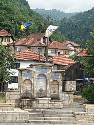 The image depicts a public fountain situated in Donja Vraca, Bosnia and Herzegovina. The fountain is constructed from light-colored stone blocks and features three arched spouts, each with a brass fixture from which water flows. Above the spouts, there are three rectangular blue plaques with Arabic inscriptions. The fountain is flanked by stone structures and is accessible via stone steps. A wooden bench is visible to the left of the fountain. Two flags are displayed prominently: one is the flag of Bosnia and Herzegovina, and the other is a white flag with a blue emblem. In the background, several houses with red-tiled roofs are visible, nestled amongst lush green hills. The sky is overcast, and the overall atmosphere suggests a tranquil, possibly historic, setting. There are no people visible in the immediate vicinity of the fountain, although there are power lines overhead.