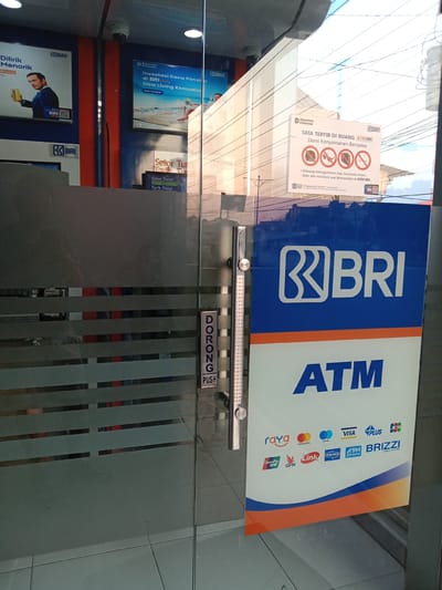 The image shows the entrance to a Bank Rakyat Indonesia (BRI) branch. The entrance is a glass door with a horizontal pull bar that reads "DORONG PUSH" vertically. On the door, a large blue and white sign displays "BRI ATM" with various payment network logos below. Inside the branch, a security camera is visible mounted on the ceiling. Posters are displayed on the walls, including one advertising "Investasi Dana Pensiun di BRI" (Pension Fund Investment at BRI) and another with a man holding a drink. A digital display near the door appears to indicate ATM transaction options. The floor is tiled. The scene is illuminated by interior lighting and natural light filtering in from outside.