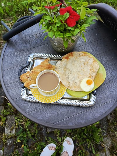 A high-angle outdoor shot shows a breakfast tray on a dark wooden table. The tray is silver and oval-shaped with ornate handles. On the tray, there are two pastries, a cup of coffee in a yellow and white patterned teacup with a matching saucer, and a green plate with a flatbread and a fried egg. Next to the tray, a pot of red petunias sits on the table. The table is surrounded by green grass and stone pavers, with the feet of a person wearing white sandals visible in the lower portion of the frame. The scene appears to be set in a garden or patio area.