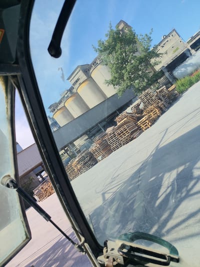 The image appears to be taken from inside a vehicle, looking out through the windshield. The scene is outdoors, with a bright blue sky and visible sunlight casting shadows. In the background, there is a large industrial complex featuring several tall, cylindrical silos with yellowish tops, and connected by elevated walkways and industrial structures. Adjacent to the silos, there are multiple stacks of wooden pallets. A large green tree is situated between the vehicle and the industrial buildings. The foreground of the image is dominated by parts of the vehicle's interior, including a steering mechanism and what looks like a windshield wiper or support arm. The ground is paved and shows some cracks. The overall impression is an industrial area with stored materials, likely a factory or processing plant. The image does not provide any specific visual cues to confirm the location within the city of Kipra, Bulgaria.