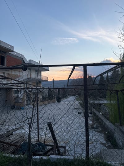 This image depicts a street scene in Strumyani, Bulgaria, viewed from an elevated perspective, likely at dusk or dawn. The foreground is dominated by a rusted chain-link fence supported by dark metal posts and horizontal bars, some of which are bent. Immediately behind the fence, a small pile of debris including corrugated metal sheets, wooden planks, and dark fabric is visible. Bare tree branches are scattered throughout the fence line. A short concrete barrier and sparse green vegetation occupy the bottom edge.
The midground features a paved street composed of light-colored, irregularly shaped stone tiles, extending into the distance. To the left of the street, a multi-story building, partially under construction, displays exposed red brick on an upper level, concrete framework, and white-painted sections. An external staircase with a white balustrade leads to a balcony featuring decorative railings. Further along the street, additional buildings are partially visible. To the right of the street, a low concrete wall separates the pavement from an area with mature, tall evergreen trees, interspersed with deciduous trees exhibiting bare branches.
In the background, rolling hills or mountains are silhouetted against a sky transitioning from clear light blue at the zenith to soft orange and pink hues near the horizon, indicative of sunrise or sunset. Wispy clouds are present in the upper sky. Utility lines run diagonally across the upper left quadrant of the frame. No individuals or active subjects are present.