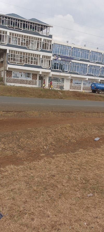 A four-story building stands prominently, featuring numerous windows and a sign reading "Queenteene Hospital." Two individuals walk in front of the building, carrying yellow containers. A water tank is parked nearby. A road separates the building from an expanse of dry, brown grass and dirt, which occupies the majority of the foreground. The sky overhead is overcast.