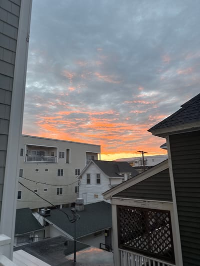 The image depicts a coastal urban scene in Hampton, United States of America, under a dramatic sky at either dawn or dusk. The sky is filled with a gradient of colors, from soft grays to vibrant oranges and pinks, suggesting illuminated clouds. In the foreground, the dark green siding and dark shingle roof of a residential building are visible on the right, partially obscuring the view. To the left, a light gray shingled wall and white trim of another building frame the scene.
Mid-ground reveals several residential structures. A large, light-colored building with horizontal siding and multiple windows stands prominently. It features balconies with white railings. Adjacent to it, a smaller white house with a dark roof is visible. Further in the background, utility poles and wires are present, indicating an established neighborhood. The rooftops are a mix of dark shingles, some appearing wet and reflecting the sky's colors. The overall impression is that of a dense residential area close to the coast. No people or specific actions are discernible in the image.
