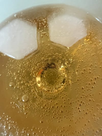 A close-up, overhead shot shows a beverage with ice cubes. The liquid is a golden-brown color and is filled with many small bubbles. Two large, irregular-shaped ice cubes are visible at the top of the frame. In the center of the image, a large bubble has formed, with a reddish-orange object partially visible inside it. The background is a light, pale blue. The scene is one of a refreshing drink.