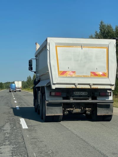 The image depicts a sunny day on a two-lane highway in Pyrizhky, Ukraine, with a clear blue sky and trees lining the right side of the road. The foreground is dominated by the rear of a white dump truck with a large, mostly white cargo bed. The truck has a license plate reading "AA 8541 XK". Two red and yellow reflective stripes are visible on the lower part of the truck's cargo bed, and there are some rust stains around the hinges. The truck is in the right lane of the highway. In the left lane, further ahead, is a blue van, followed by a white semi-trailer truck. The highway has white lane markings.