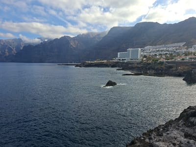 A wide-angle, outdoor, daytime shot captures a coastal view of Puerto de Santiago, Spain, featuring rugged cliffs, the ocean, and a resort. The foreground is dominated by dark, rocky terrain. The middle ground consists of the deep blue ocean, with a small rock formation emerging from the water near the center and waves lapping at its base. A rocky coastline with a pier extends into the water on the right side of the frame. In the background, large, imposing, dark-colored cliffs rise dramatically. At the base of the cliffs, a white, multi-story hotel with many windows and balconies is visible, along with a densely populated resort complex featuring numerous white buildings, roads, and palm trees. The sky above is a mix of blue and white clouds, with sunlight casting shadows and highlights on the landscape. There are no people visible in the scene.