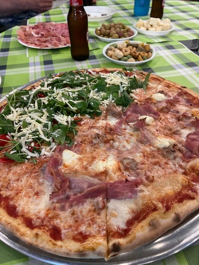 In Toritto, Italy, a close-up shot captures a large, halved pizza with two distinct toppings. One half is generously covered with fresh arugula and shredded cheese, while the other features prosciutto and dollops of white cheese. The pizza rests on a silver platter on a green and white checkered tablecloth. In the background, several small bowls contain olives, cheese cubes, and crackers. A brown bottle of beer and a plate of thinly sliced cured meats are also visible, suggesting a meal shared amongst people whose arms and hands are partially visible, indicating a social gathering. The scene appears to be an outdoor or casual dining setting.