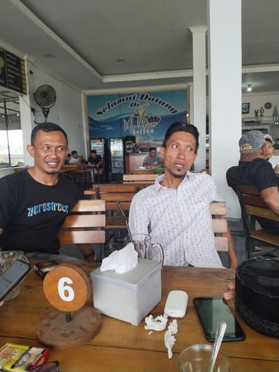 This media file shows a group of men sitting at tables in what appears to be a cafe or restaurant. Two men are in the foreground, one on the left wearing a black t-shirt with "aerostreet" printed on it, and another on the right wearing a white patterned shirt. Both men are looking away from the camera, with the man in the black shirt smiling. In the background, other patrons can be seen at tables, and there's a large mural on the wall with the text "Selamat Datang Di Mall Coffee."

Objects on the table in the foreground include a black smartphone, a tissue box, a wooden table number with the number "6" on it, cigarette packs, and glasses. The setting seems to be indoors, possibly with natural light coming from the left, suggesting it's daytime. The overall mood appears relaxed and casual. The text on the mural and the table number provide clues about the location being a cafe. The presence of food items like "KENTANG GORENG," "NUGGET," "SOSIS," and "TEMPE GORENG" on a menu board in the background further confirms the establishment serves food. A fan is visible on the wall, indicating a warm climate. The man on the right is wearing a black baseball cap with text that includes "HAPPY BLACK PLAND," and another man in the background wears a similar cap.