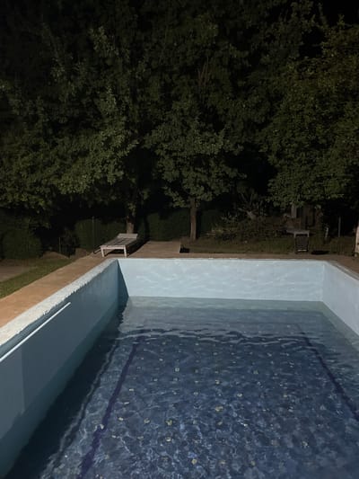 The image depicts an outdoor swimming pool at night. The pool is partially filled with water, reflecting the surrounding dark trees and a clear night sky. The pool features a light blue interior with a dark blue tiled bottom marked by a purple line. A light-colored lounge chair is visible on the paved area next to the pool. In the background, dense foliage and trees create a natural, somewhat overgrown setting. The scene is dimly lit, suggesting it is either late at night or the area is not brightly illuminated. There are no people or visible signs of recent activity. The environment appears to be a private residential area or a small establishment in Машат, Kazakhstan, characterized by its lush vegetation and the presence of a swimming facility.