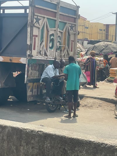 This image depicts an outdoor urban environment in Awjaw, Nigeria, characterized by commercial activity. In the midground, three adult males are positioned around a dark-colored motorcycle and the rear of a large truck. One man, wearing a light-colored shirt and dark trousers, is seated on the motorcycle, holding a white smartphone with a white cable, and is looking towards the other two. A second man, wearing a teal polo shirt and dark shorts, stands facing the seated man, with his back to the viewer. A third man, partially obscured and wearing a light brown top, stands between the two. The rear of the truck, painted white with geometric patterns in red, green, and light blue, features the numbers "5" and "6," and partial text "ZAT KT" is visible below. The truck's body is dark blue with a light blue tarp.
To the right of the group and extending into the background, a busy market scene unfolds. Large piles of white sacks, indicative of bulk goods, are visible, many covered by dark tarpaulins that form makeshift shelters. A woman with a light head covering, dressed in a vertically striped garment (blue, white, brown, red), is observed walking or standing near these piles, carrying a pink bag. In the far background, a light yellow building under construction, featuring scaffolding, is visible under a hazy sky. The ground in the midground is an uneven dirt and asphalt surface, partially separated by a concrete barrier in the foreground.