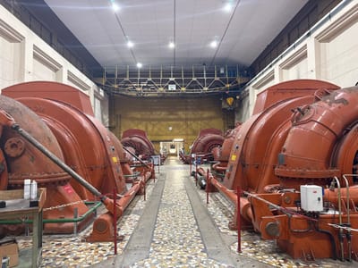 The image depicts the interior of an industrial power generation facility, likely a hydroelectric powerhouse, located in Pellafol, France. The scene is dominated by two parallel rows of large, rust-orange industrial machines, presumed to be turbines or generators, extending from the foreground into the background. Each machine features robust, curved metal casings with numerous bolted panels, pipes, and valves. The closest machine on the left has "THOM" visible on its casing, and a diagonal metal pipe runs across its front. The closest machine on the right features a small, white control box with red accents mounted on its side.

A central aisle with a patterned mosaic tile floor, consisting of various earthy tones (brown, beige, orange, grey), separates the two rows of machinery. Metal grates or channels are embedded in the floor along the bases of the machines. Red metal posts with chains serve as safety barriers along the pathways adjacent to the machines. Various pipes, some painted green, are visible connecting to the machinery at ground level.

The facility features light-colored, possibly cream-colored, walls with recessed paneling. The ceiling is flat and paneled, illuminated by multiple recessed artificial light fixtures that provide bright, even light throughout the space. Towards the back and upper section of the room, a yellow gantry crane structure is visible, along with elevated walkways or support beams. A doorway or opening is discernible in the back wall, at the end of the central aisle. No individuals are present in the scene, and no specific actions or interactions are depicted. The machinery shows signs of use, including some surface discoloration and minor rust. A dark workbench with several white spray cans and a cardboard box is visible in the extreme left foreground.