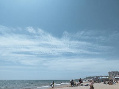 The image depicts a beach scene with several people enjoying a sunny day. In the foreground, people are relaxing on lounge chairs and towels on the sand, some under umbrellas. Two individuals stand near the water's edge, one bending down. The ocean is visible in the midground with gentle waves. In the background, several buildings, possibly hotels or apartments, are situated along the coastline, suggesting an urban or resort area. The sky is clear blue with wispy clouds. The overall impression is a lively beach environment. The scene is consistent with a summer day at the beach in Rota, Spain, a coastal city known for its beaches and seaside resorts.