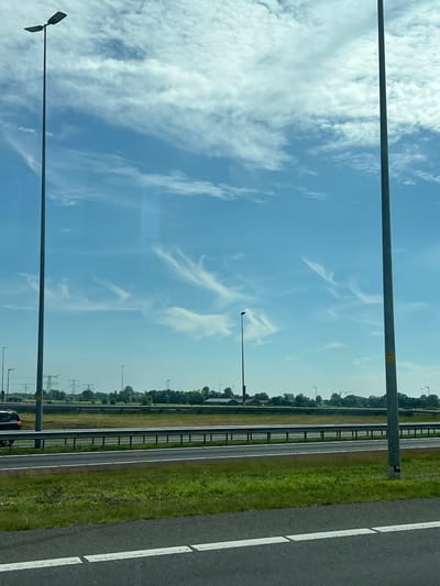The image depicts a highway scene under a bright, partially cloudy sky. In the foreground, a paved road with white lane markings is visible. To the right, a tall, dark grey lamppost stands vertically, with a yellow sticker affixed to its base. Beyond the road, a grassy median separates lanes, and another section of highway with a metal guardrail is present. A dark-colored car is visible on the left side of the highway. In the background, a line of trees forms a distant horizon, interspersed with utility poles and what appear to be electrical transmission towers. Several other lampposts are scattered across the landscape, indicating the presence of a well-lit roadway. The sky is a clear blue with scattered white, wispy clouds. There are no people visible in the image. The scene suggests a location on the outskirts of a city, possibly an industrial or commercial area, given the infrastructure. Leeuwarden, Netherlands, is a city with a network of roads and similar infrastructure, making this a plausible setting.