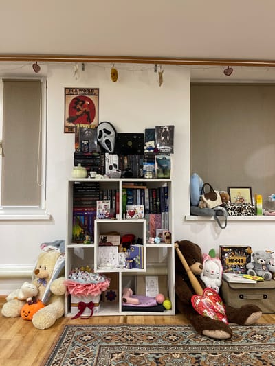 The image shows a room, likely a child's bedroom or playroom, with a white shelving unit filled with books and stuffed animals. The room is decorated with fairy lights and various whimsical items. On the floor in front of the shelves are several large stuffed bears and a plush pumpkin. To the right of the shelves, on a windowsill, are more stuffed animals, a framed picture, and other decorative items. The overall impression is one of a cozy and imaginative space.