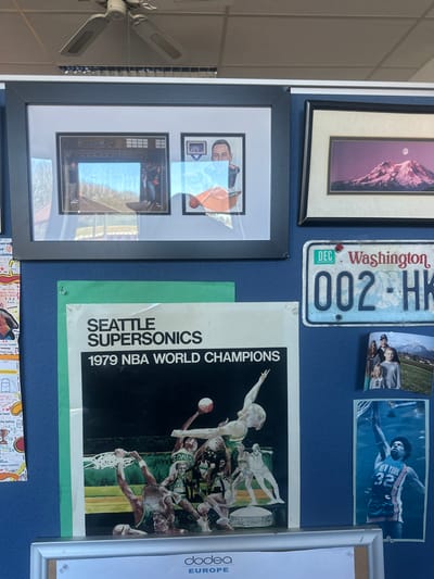 The image shows a wall in a room in Münchweiler an der Alsenz, Germany, with various items pinned up on it. The wall is painted a dark blue color.  The largest item on the wall is a poster of the Seattle Supersonics, the 1979 NBA world champions. There are framed pictures of a man playing basketball and a mountain landscape, as well as a Washington State license plate. There is also a photo of a family, a photo of a basketball player, and a small, framed picture that reads "dodea EUROPE." The ceiling has a white fan.