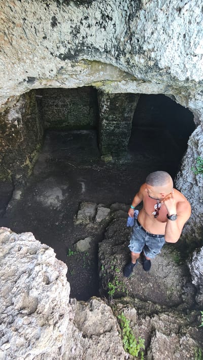 A man stands at the edge of a stone pit or hole in the ground. The location is Kamen Bryag, Bulgaria. The man is shirtless, wearing denim shorts, dark shoes, a watch, and sunglasses hanging from his neck. He holds a light-colored cloth in his hand. The pit is surrounded by rough, white and gray rocks with some green vegetation. The interior of the pit is dark and appears to have carved stone walls. The perspective is from above.