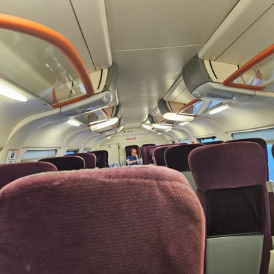The interior of a train car. Rows of plum-colored seats are visible on either side of a central aisle. An adult male wearing a blue shirt is seated in one of the seats in the middle of the train car. Above the seats on each side of the aisle are handrails with transparent panels. Windows are along the upper portion of the walls of the train car. The light fixtures appear to be linear fluorescent lamps. It's unlikely this photo was taken in El Mansouria, Morocco.