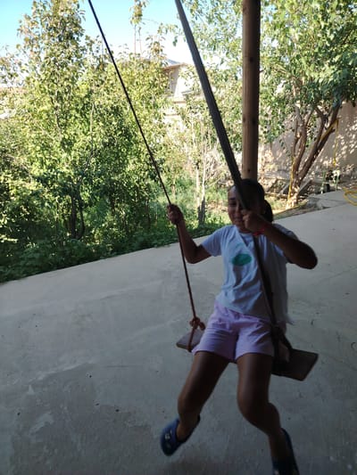 A young girl with dark hair is swinging on a wooden swing suspended from a rope. She is wearing a light-colored t-shirt with a green design and light pink shorts. Her feet are clad in blue slip-on shoes. She is holding onto the swing's ropes and appears to be in motion, with her body angled slightly. The swing is set in an outdoor area with a concrete ground. Behind the girl, there are numerous trees with green foliage, creating a natural backdrop. A wooden pillar is visible to the right of the girl, supporting an overhead structure which is partially obscured by the trees. In the distance, a building with a light-colored roof is visible amongst the trees. The environment suggests a residential backyard or a park area. The scene is brightly lit by daylight, casting shadows on the concrete surface.