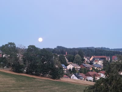 The image depicts a twilight scene in the village of St. Märgen, Germany. A bright, full moon is visible in the clear, pale blue sky in the upper left quadrant. The foreground consists of a gently sloping, dry, mown field. Beyond the field, a densely wooded area with various trees, some with dark green foliage, lines the middle ground. Nestled within the trees and extending into the background are numerous residential houses, characterized by pitched roofs in shades of red, brown, and gray, with visible windows and balconies. Some houses are multi-story structures. A tall, reddish-brown brick chimney protrudes from behind the trees in the central background. The overall impression is a peaceful, rural residential area at dusk, with the moon ascending. No people are visible in the image.