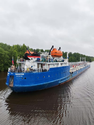 An outdoor, mid-range, eye-level, vertical, full shot shows a blue and white cargo ship named "BANATOV" sailing on a body of water, likely a river. The ship is seen from its port bow to its mid-section. The ship's bow is blue, and the superstructure is white with blue accents. There are two orange life rafts visible on the upper deck, one on the port side and another slightly further back on the starboard side. A Russian flag is flying from a pole on the port side of the bow. The ship's name, "BANATOV," and the city "VOLGOGRAD" are written in Cyrillic on the blue hull near the bow. The ship appears to be moving through calm, dark water. The background consists of a dense line of green trees on the left bank of the river, suggesting a natural, possibly rural, environment. The sky is overcast and a pale gray. The scene is static, with no people or overt actions visible on the ship. The image was taken in Volokov Most, Russia, a location characterized by waterways and surrounding forests.