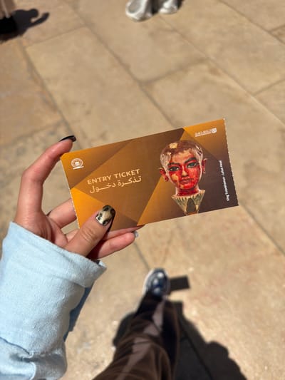 In Kafrat Nassar, Egypt, a hand with patterned fingernails holds an entry ticket. The ticket features an image of a red-faced pharaonic head, possibly King Tutankhamun's Lotus Head, and text in English and Arabic. The background shows a sunlit, tiled ground and shadows cast by people.