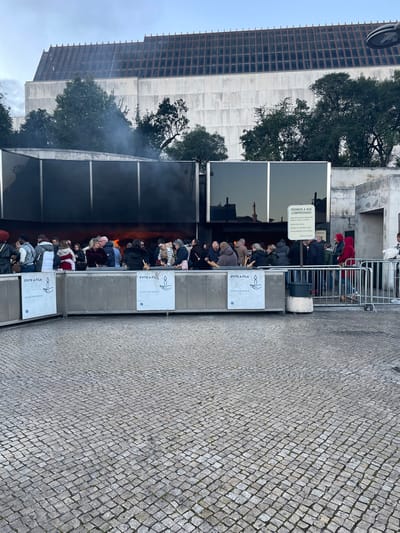A gathering of people are standing behind metal barriers. Smoke fills the background. The barriers are marked with signs in a foreign language and candle icons. A large building is visible on the left with trees positioned between it and the crowd. The ground appears to be stone. Some individuals stand near a sign that says "PEDIMOS A SUA COMPREENSÃO." The scene suggests an event or place where candles are burned, potentially a memorial or religious site.
