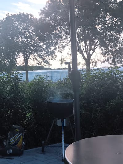 This image captures an outdoor scene, likely in Almere, Netherlands. The focal point is a charcoal grill with a bag of charcoal briquettes beside it, indicating a barbecue is either in progress or about to begin. Smoke is rising from the grill, suggesting it is hot.
Behind the grill, a dense hedge of green foliage forms a natural backdrop. Further in the distance, a large, modern greenhouse structure with a light blue roof is visible, hinting at an agricultural or horticultural setting. Silhouetted against a bright, possibly early evening sky with scattered clouds are the dark shapes of large trees. The lighting suggests it's either late afternoon or early evening, with the sun low in the sky.
A dark grey, round tabletop is partially visible in the foreground, suggesting a dining or seating area. A tall, dark pole, likely part of a patio umbrella, stands vertically between the grill and the viewer, obscuring a small portion of the scene.
The bag of charcoal briquettes has text in Dutch and possibly other languages. Visible text includes "Houtskool Briketten", "Holzkohle Briketts", and "Briquettes de Charbon de Bois". There is also a logo that appears to be "FHM".
The overall mood is relaxed and suggests a pleasant outdoor gathering. There are no people visible in the image.