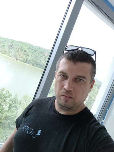 The image is a selfie taken by a man standing indoors next to a large window. The man is wearing a black t-shirt with a light blue graphic on the left chest. Sunglasses are perched on top of his head. The window overlooks a body of water, possibly Lake Mácha, and a densely wooded hillside. The man's facial features are visible, and he appears to be looking at the camera. The overall setting suggests a scenic outdoor view from an indoor location.