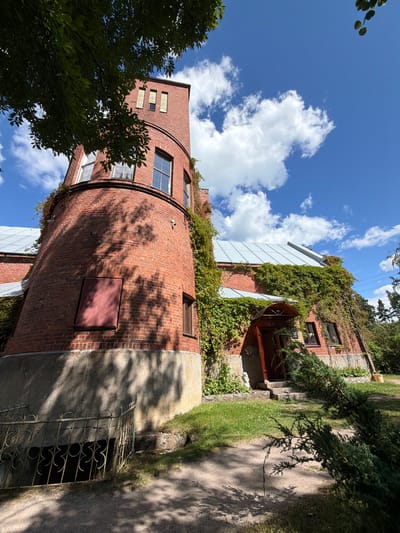 The image shows a red brick building with a rounded tower and a sloped metal roof, partially covered in green ivy. The building is situated in a park-like setting with green grass, trees, and a blue sky with white clouds. The facade of the building features several windows, an arched entrance with a wooden door and a small awning, and a dark red rectangular panel on the tower. A metal fence with decorative elements encloses a dark opening in the ground near the tower. The scene is illuminated by sunlight, casting shadows from trees onto the building and ground. The building is located in Sevastyanovo, Russia.