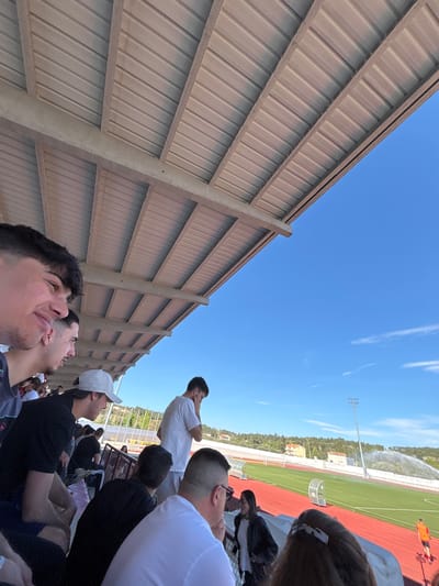 The image depicts a group of people at a sports stadium, likely in Valpaços, Portugal, based on the architecture and landscaping. The stadium has a running track and a field, with a watering system active in the background. People are sitting in the stands, some looking up towards the field. The stands are constructed with metal siding and concrete supports. The sky is clear and blue.