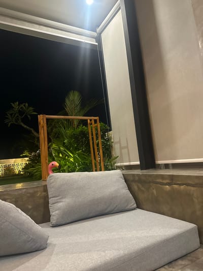 The image displays an outdoor or semi-outdoor seating area at night in Tumbak Bayuh, Indonesia. In the foreground, a grey, upholstered lounge sofa with multiple cushions is present, constructed upon a concrete base. To the left and slightly behind the sofa, the pink head of a plastic flamingo lawn ornament is visible amongst dense green tropical foliage. Behind the flamingo, a freestanding wooden frame structure is positioned amidst the vegetation, which includes palm fronds. In the far background, a white, geometrically patterned wall or fence is faintly illuminated. The sky is dark, indicating nighttime. Overhead, a bright, recessed LED spotlight casts light downward onto the scene. Adjacent to the light, a white roller blind mechanism is attached to the ceiling. A light-colored plastered wall and a dark vertical support beam of a building structure are visible on the right side of the frame. No individuals are present in the scene.
